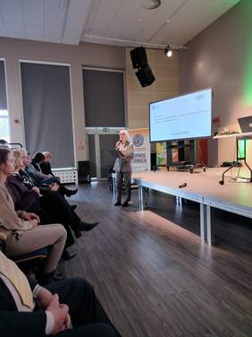 Pokaż zdjęcie: Po prawej stronie stoi kobieta, która przemawia do mikrofonu. Jest elegancko ubrana. Po lewej stronie siedzą na krzesłach uczestnicy konferencji. 