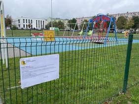 Pokaż zdjęcie: Na ogrodzeniu znajduje się plakat informacyjny. Napisane jest na nim, że plac zabaw dla dzieci został zrealizowany w ramach programu „Dostępna Przestrzeń Publiczna”. Plakat zawiera także inne informacje. W tle widać huśtawki, karuzele i inne urządzenia dla dzieci. 