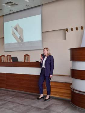 Pokaż zdjęcie: Przed stołem konferencyjnym stoi z mikrofonem w ręku kobieta, blondynka w ciemnym garniturze.
