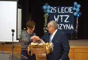 Pokaż zdjęcie: Kobieta wręcza mężczyźnie kosz upominkowy. W tle widoczny jest napis: „25-lecie WTZ w Rydzynie” udekorowany niebieskimi balonami.