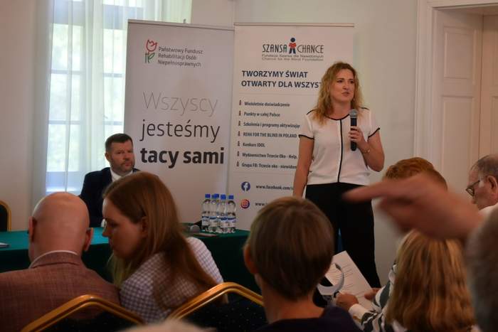 Pokaż zdjęcie: Kobieta z mikrofonem stoi w sali konferencyjnej na tle baneru Fundacji Szansa dla Niewidomych. Wystąpieniu przysłuchuje się grupa kilku osób. Za stolikiem przykrytym zielonym płótnem siedzi mężczyzna, obok którego ustawiony jest baner PFRON.