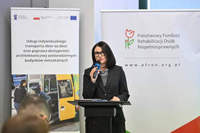 Pokaż zdjęcie: brunetka w czarnej marynarce i okularach, stoi przy mównicy. Trzyma w ręku mikrofon. W tle plansze projektu z logotypami 