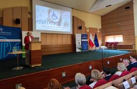 Pokaż zdjęcie: Kobieta w czerwonej marynarce przemawia przy mównicy podczas konferencji w przestronnej sali wykładowej. Na ekranie za nią widnieje slajd z napisem: ‘KONFERENCJA – Prawne i pozaprawne aspekty zatrudniania osób z niepełnosprawnościami’. Obok znajdują się banery organizatorów, a także flagi Polski i Unii Europejskiej.