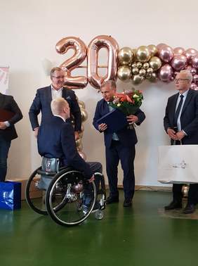 Pokaż zdjęcie: Mężczyzna ubrany w granatowy garnitur trzyma w ręku duży grawer i kwiaty. Mówi do siedzącego przednim na wózku mężczyzny również ubranego w garnitur. Nad nimi na ścianie wisi nadmuchana z srebrnego balonu cyfra 20.