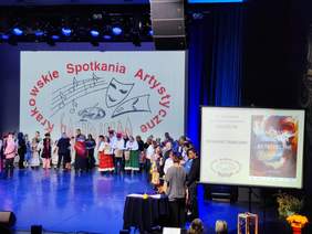 Pokaż zdjęcie: Scena pełna uczestników Spotkań Artystycznych. Za uczestnikami w strojach regionalnych widoczne logo wydarzenia. Obok sceny plakat z logo wydarzenia i nazwą kategorii konkursowej: Konkurs taneczny