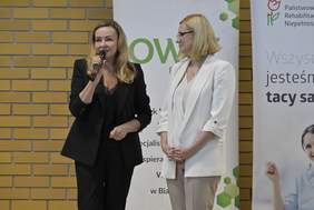 Pokaż zdjęcie: Dwie kobiety stoją na scenie na tle żółtej ceglanej ściany. Jedna z nich, ubrana w czarny żakiet, bluzkę i spodnie, trzyma mikrofon i przemawia. Obok niej stoi druga kobieta w jasnym kostiumie, uważnie słuchając. Nad nimi widoczne są banery z logo PFRON i OWiT.