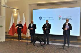Pokaż zdjęcie: Czterech mężczyzn stoi na pierwszym planie. Jeden z nich przemawia. Z lewej strony stoi tłumaczka języka migowego. W tle flagi narodowe i ekran z logiem Ministerstwa i PFRON-u.