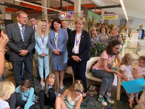 Pokaż zdjęcie: We wnętrzu galerii cztery osoby w eleganckich strojach stoją wśród dzieci malujacych obrazki. Dzieci siedzą na podłodze. Z tyłu wiele postaci przyglądajacych się.