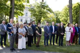 Pokaż zdjęcie: Grupa kilkunastu osób zwrócona w jednym kierunku. W tle fragment parku i okazały budynek