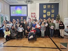 Grupa osób pozuje do fotografii. Uczestnicy trzymają w rękach otrzymane dyplomy i nagrody. Osoby z niepełnosprawnościami, w tym na wózkach, stoją i siedzą w pierwszym rzędzie
