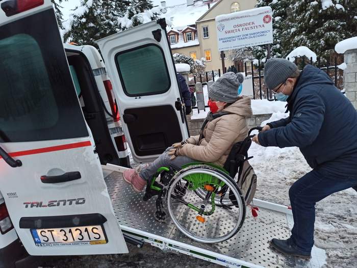 Pokaż zdjęcie: widok tyłu busa z otwartymi drzwiami i wysuniętą platformą po której wjeżdża dziewczyna na wózu inwalidzkim z pomocą mężczyzny w tle ośnieżone choinki i budynek ośrodka