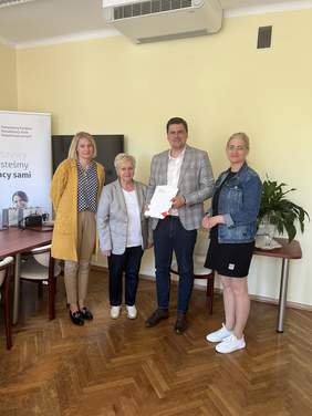 Pokaż zdjęcie: Jeden mężczyzna i trzy kobiety stoją na tle ściany z banerem reklamującym PFRON. Mężczyzna trzyma w rękach teczki. Wszyscy są uśmiechnięci