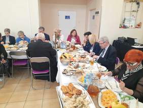 Pokaż zdjęcie: Przy stołach siedzą zaproszeni goście. Wszyscy jedzą świąteczne potrawy. Stoły są zastawione różnymi smakołykami: pierogami, karpiem, śledziami, domowymi wypiekami. 