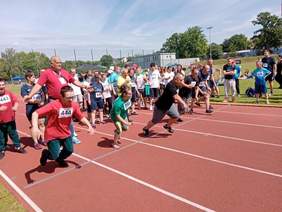 Pokaż zdjęcie: Po czerwonej bieżni biegnie kilku sportowców z niepełnosprawnością. W tle widać także innych zawodników. 