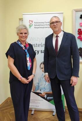 Pokaż zdjęcie: Uśmiechnięta kobieta i mężczyzna ubrani elegancko znajdują się w pomieszczeniu z jasnymi ścianami. Za nimi baner z logo PFRON oraz kolorowa praca plastyczna.