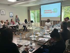 Pokaż zdjęcie: Sala konferencyjna. Uczestnicy siedzą przy stolikach i słuchają prelegenta. Na ekranie wyświetlane są procedury programu PFRON.