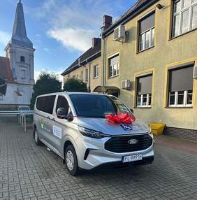 Pokaż zdjęcie: Srebrny bus przyozdobiony czerwoną kokardą na tle znajduje się budtynek o żółtej elewacji, a za nim widoczny jest kościół z wysoką wieżą