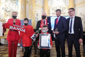 Pokaż zdjęcie: Na scenie stoi kobieta, trzech mężczyzn i dwójka dzieci. W rękach trzymają kwiat, statuetki i dyplomy. Kobieta z dzieckiem trzymają rozciągnięty szalik z napisem "futbolowa banda"