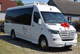 Pokaż zdjęcie: Biały autobus z napisem „Gmina Szydłowo”, naklejką informującą, że pojazd przystosowany jest do przewozu osób z niepełnosprawnością oraz czerwoną kokardą przymocowaną z przodu, na masce. W tle dwa samochody osobowe oraz zabudowania mieszkalne.