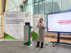 Pokaż zdjęcie: Osoba prowadzi prezentację przy mównicy podczas wydarzenia PFRON. W tle stoją roll‑upy informacyjne oraz duży ekran z tytułem dotyczącym wyrównywania różnic między regionami.