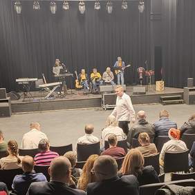 Pokaż zdjęcie: Na zdjęciu widać scenę w sali teatralnej lub koncertowej. Na scenie znajduje się kilku muzyków przygotowujących się do występu lub w trakcie próby. Niektórzy grają na gitarach, a jeden z nich siedzi przy keyboardzie i śpiewa do mikrofonu. W tle widoczne są dodatkowe instrumenty, takie jak perkusja oraz bębny.  Publiczność siedzi w rzędach krzeseł przed sceną, a część osób jeszcze zajmuje miejsca. Jeden mężczyzna w białej koszuli przechodzi na pierwszym planie. Widzowie są ubrani różnorodnie, a niektóre osoby mają na głowach mikołajkowe czapki, co sugeruje, że wydarzenie może być związane z okresem świątecznym. Atmosfera zdjęcia wskazuje na spokojne przygotowania do koncertu lub występu.