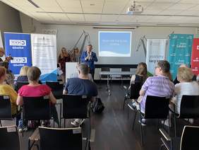 Pokaż zdjęcie: Na Sali konferencyjnej na krzesłach siedzą ludzie. Przed nimi  stoi mężczyzna ubrany w niebieską marynarkę i białą koszulę trzyma w ręku mikrofon. Za nim prezentacja z napisem reha for the blind Gdańsk.
