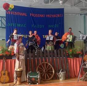 Pokaż zdjęcie: Na pierwszym planie scena, na której sześć osób ubranych w bogato zdobione stroje tańczy i śpiewa. Za nimi napis VII Festiwal Piosenki Przyjaznej ,,Śpiewać każdy może’’ na granatowym tle