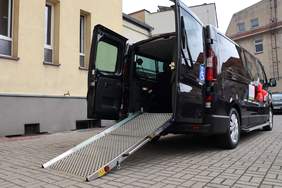 Pokaż zdjęcie: Bus z dwoma naklejkami informującymi, że pojazd przystosowany jest do przewozu osób z niepełnosprawnością oraz dofinansowany ze środków PFRON. Bus ma otwarte tylne drzwi oraz wyciągniętą rampę. Pojazd stoi na dziedzińcu pomiędzy kilkoma budynkami.