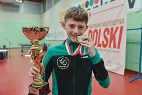 Pokaż zdjęcie: Młody chłopak z medalem w zębach i złotym trofeum w rękach