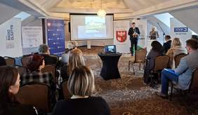 Pokaż zdjęcie: Kilkanaście osób siedzi na krzesłach w sali konferencyjnej. Przed nimi stoi kobieta z mikrofonem oraz mężczyzna w garniturze. Za nimi prezentacja wyświetlona na ekranie oraz banery: PFON, TVP3, Radio Olsztyn, Wojewody Warmińsko-Mazurskiego oraz Marszałka Województwa Warmińsko-Mazurskiego