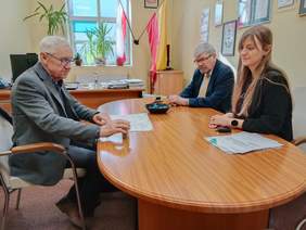 Pokaż zdjęcie: Przy podłużnym stole siedzi kobieta i dwóch mężczyzn. Podpisują umowę. Na stole leżą różne dokumenty, pieczątki, długopisy. 