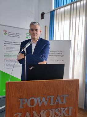 Pokaż zdjęcie: Przy mównicy stoi wysoki mężczyzna, ubrany w ciemną marynarkę, ma skrzyżowane ręce.