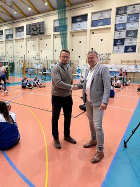Pokaż zdjęcie: Dwóch mężczyzn stoi na boisku sportowym i uściska sobie dłonie. Po lewej stronie jest mężczyzna z okularami, ubrany w szary sweter i ciemne spodnie. Mężczyzna po prawej ma na sobie jasnoszarą marynarkę, białą koszulę i jasne jeansy. Obaj patrzą na siebie z uśmiechem. W tle, na pomarańczowej podłodze boiska sportowego, widać inne osoby siedzące na ziemi, a także siatkę. Na ścianie za nimi widoczne są tablice reklamowe sponsorów, w tym Grupa Azoty. Scena sugeruje moment oficjalnego powitania lub podziękowania podczas wydarzenia sportowego.