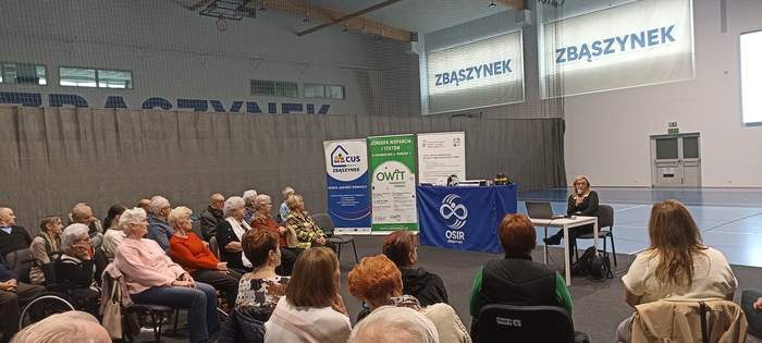 Pokaż zdjęcie: Widoczni uczestnicy spotkania siedzący na krzesłach, a przed nimi rollupy i prelegentka z mikrofonem w ręku