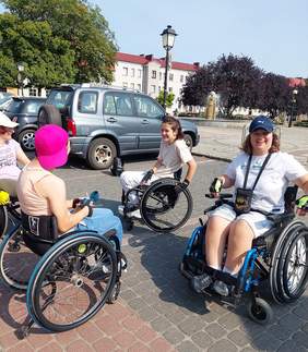 Pokaż zdjęcie: Cztery kobiety poruszające się na wózku znajdują się na parkingu. W tle: samochody osobowe, fontanna, budynek mieszkalny, latarnie oraz drzewa liściaste.