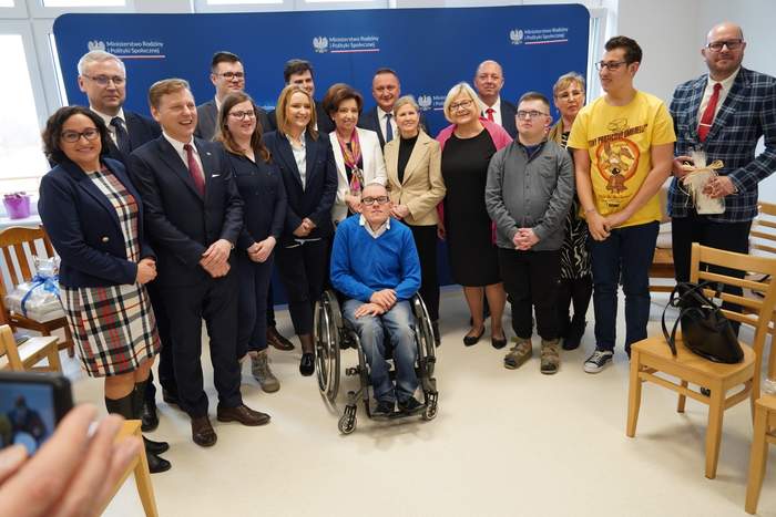 Pokaż zdjęcie: Kilkanaście osób stoi w dwóch rzędach na tle banneru reklamującego Ministerstwo Rodziny i Polityki Społecznej. Wszyscy są ubrani odświętnie