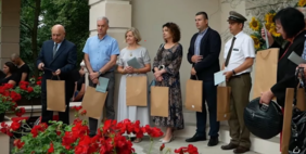 Pokaż zdjęcie: Zdjęcie na którym widać grupę siedmiu osób stojących w półkolu trzymających w ręku torebki prezentowe, zdjęcie zrobione przed wejściem