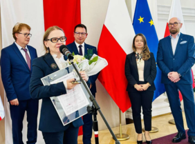 Pokaż zdjęcie: Przy mikrofonie stoi laureatka konkursu, w ręku trzyma kwiaty i statuetkę. Z tyłu stoją pozostali laureaci.