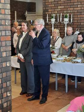 Pokaż zdjęcie: Na środku sali stoi kobieta i mężczyzna. Pan przemawia do mikrofonu. W tle widać innych ludzi i świątecznie zastawione stoły. 