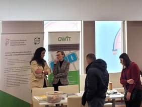 Pokaż zdjęcie: Stoiska informacyjne wystawców podczas Targów Pracy. Uczestnicy podchodzą do stoisk i przeglądają materiały informacyjne. Za stoiskami stoją rollupy, na których są logotypy m.in.  PFRON i OWiT. 