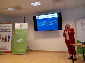 Pokaż zdjęcie: Kobieta blondynka w czerwonym garniturze prowadzi wykład podczas konferencji.