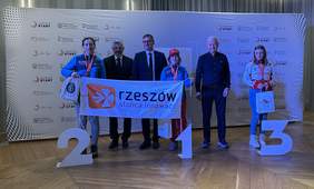 3 zawodniczki klubu, pod nimi duże cyfry symbolizujące pozycję. od lewej 2. 1, 3. Pomiędzy Paniami 3 Panów. Trzymają baner z napisem Rzeszów stolica innowacji. Za nimi duży baner z różnymi logami w tym PFRON