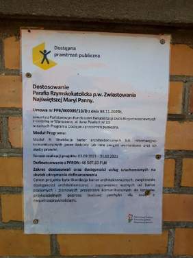 Pokaż zdjęcie: Na murze z miodowej cegły wisi biała tablica z informacją o projekcie Dostępna przestrzeń publiczna.