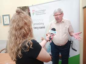 Pokaż zdjęcie: Dyrektor Łódzkiego Oddziału PFRON udziela wywiadu dziennikarce telewizji TOYA. Widać także fragment kamery. W tle widać równieź baner Łódzkiego Oddziału PFRON. 