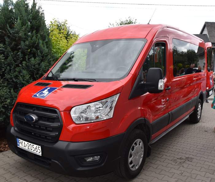 Pokaż zdjęcie: Czerwony bus z naklejką informującą, że pojazd przystosowany jest do przewozu osób z niepełnosprawnością. W bocznej szybie samochodu odbija się jak w lusterku grupa kilku zgromadzonych w pobliżu osób. W tle drzewa i krzewy oraz fragment budynku.
