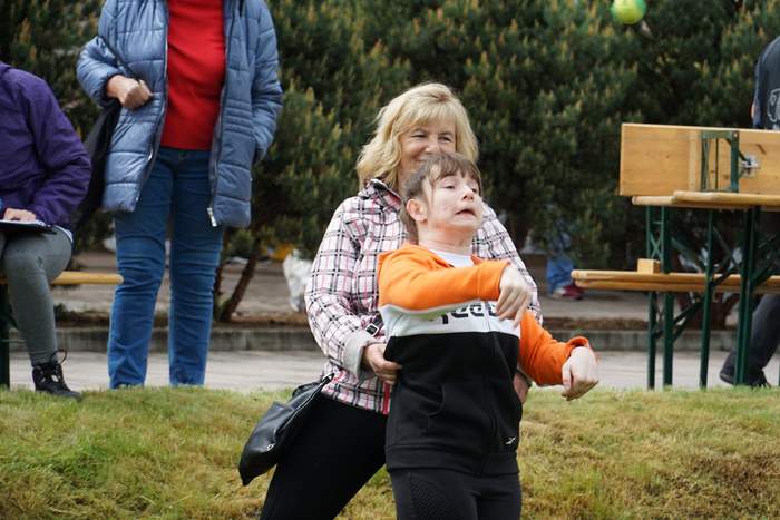 Pokaż zdjęcie: Dziewczynka bierze udział w konkurencji sportowej, podtrzymywana przez kobietę rzuca małą piłką