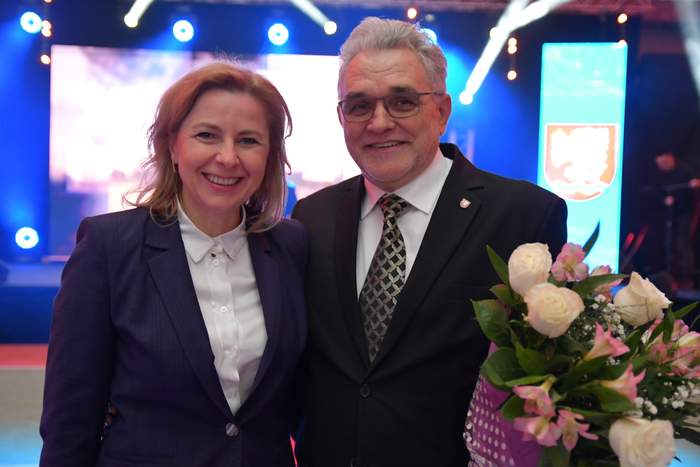 Pokaż zdjęcie: Dwie osoby, mężczyzna i kobieta, ubrani w eleganckie garnitury, mężczyzna trzyma kwiaty w ręku, obejmuje kobietę ramieniem.