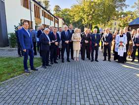 Pokaż zdjęcie: Grupa osób w eleganckich strojach stoi na brukowanym placu przed jasnym budynkiem; w centrum kobieta w beżowym płaszczu, obok mężczyźni w garniturach i duchowny w białej stule z kaszubskim haftem, w tle drzewa i zabudowania.