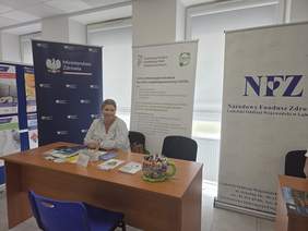 Pokaż zdjęcie: Stoisko informacyjne PFRON podczas wydarzenia. Przy biurku siedzi pracownik, w tle widoczne są roll-upy PFRON, CIDON, NFZ i Ministerstwa Zdrowia oraz materiały informacyjne.