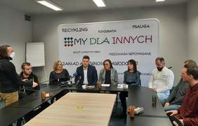 Pokaż zdjęcie: osiem osób siedzi przy stole po lewej przed nimi stoi mężczyzna w maseczce w dłoni trzyma mikrofon, za mini baner z logo stowarzyszenia my dla innych.
